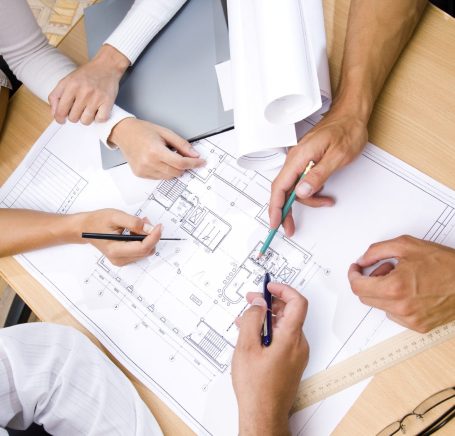 Tisch mit Architektenplan und Händen die darauf zeigen oder schreiben. Stellt eine Baubesprechung dar.